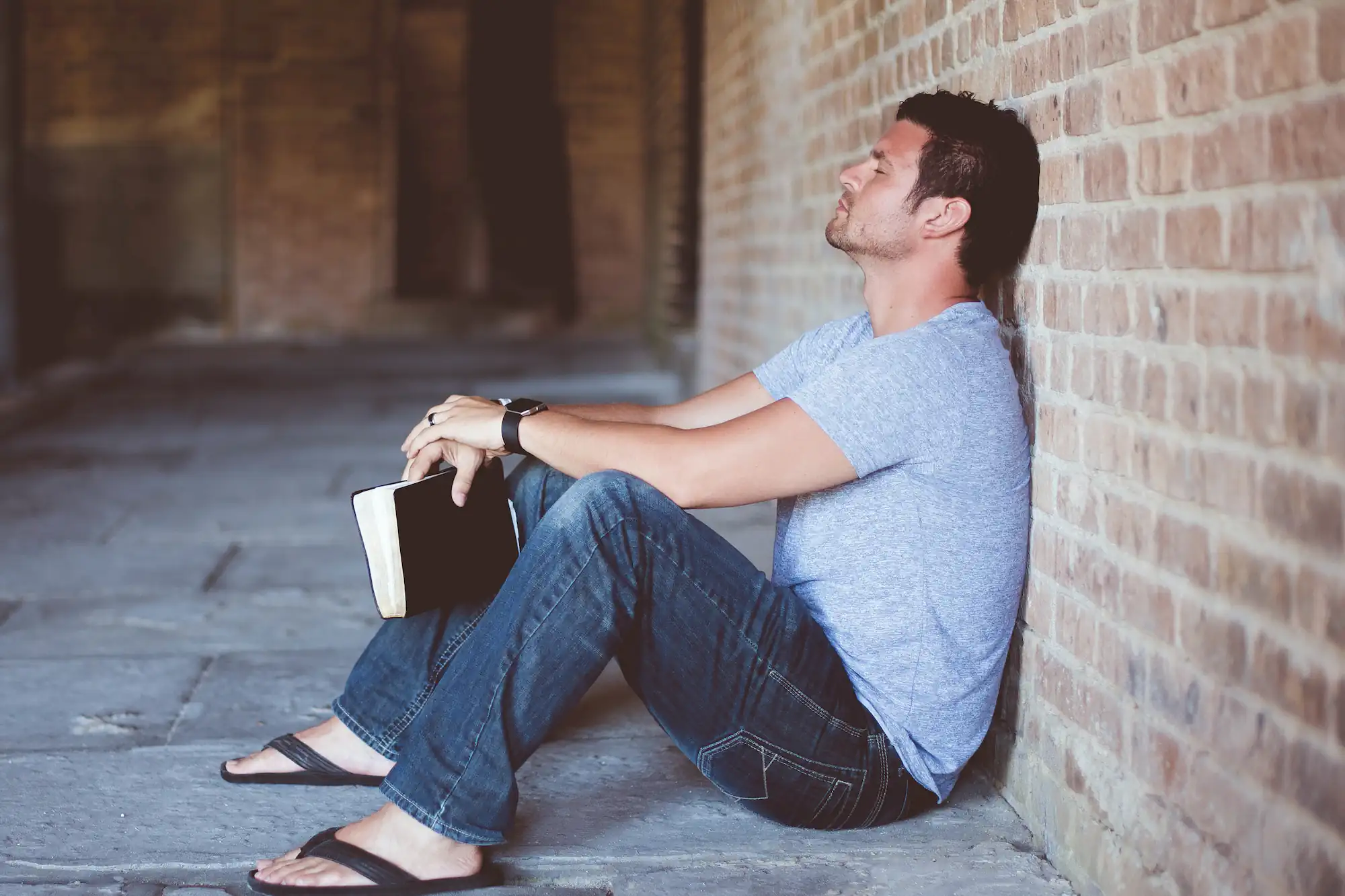 Man sitting against a brick wall holding a Bible